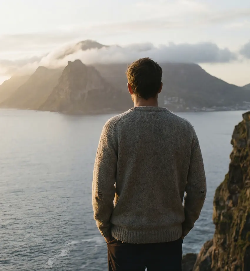Una persona con un jersey gris está de pie en un acantilado rocoso, contemplando el océano con las montañas al fondo, bajo un cielo nublado al atardecer.